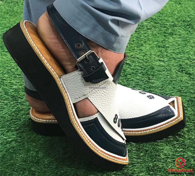May include: White and blue leather sandals with a black strap and buckle. The sandals have a thick black sole and a white stitched trim.