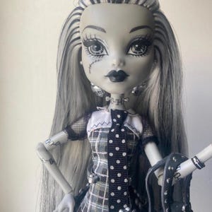 Puede incluir: Una muñeca de piel gris claro, con cabello largo y liso con mechas negras y blancas, y una cicatriz cosida en la mejilla derecha. La muñeca viste un vestido de cuadros con cuello blanco, corbata negra de lunares, cinturón y bolso con tachuelas.