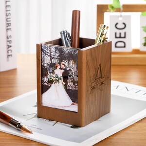 May include: A wooden pen holder with a photo of a couple in front of a wrought iron gate. The pen holder has a personalized initial and name. Several pens are inside the holder, including a silver pen and a gold pen.