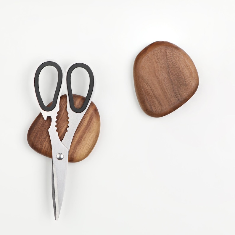 May include: A pair of stainless steel scissors with black handles, resting on a small, irregularly shaped wooden base. A second, similar wooden piece is positioned nearby. The scissors have a serrated edge and a silver rivet.