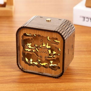 May include: A wooden, cube-shaped night light featuring a carved design of a cartoon character, trees, and a crescent moon. The light shines through the cutouts, creating a warm glow. The night light is made of light brown wood.