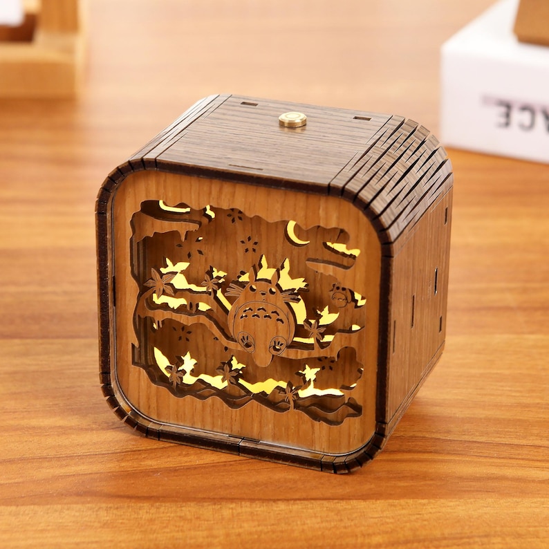 May include: A wooden, cube-shaped night light featuring a carved design of a cartoon character, trees, and a crescent moon. The light shines through the cutouts, creating a warm glow. The night light is made of light brown wood.
