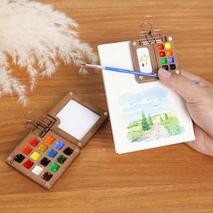 Pode incluir: Duas paletas de aquarela de madeira com uma variedade de cores vibrantes. Uma paleta está aberta, exibindo as tintas, enquanto a outra está presa a um pequeno caderno de esboços com um clipe. Um pincel repousa sobre o caderno, que apresenta uma pintura em aquarela.