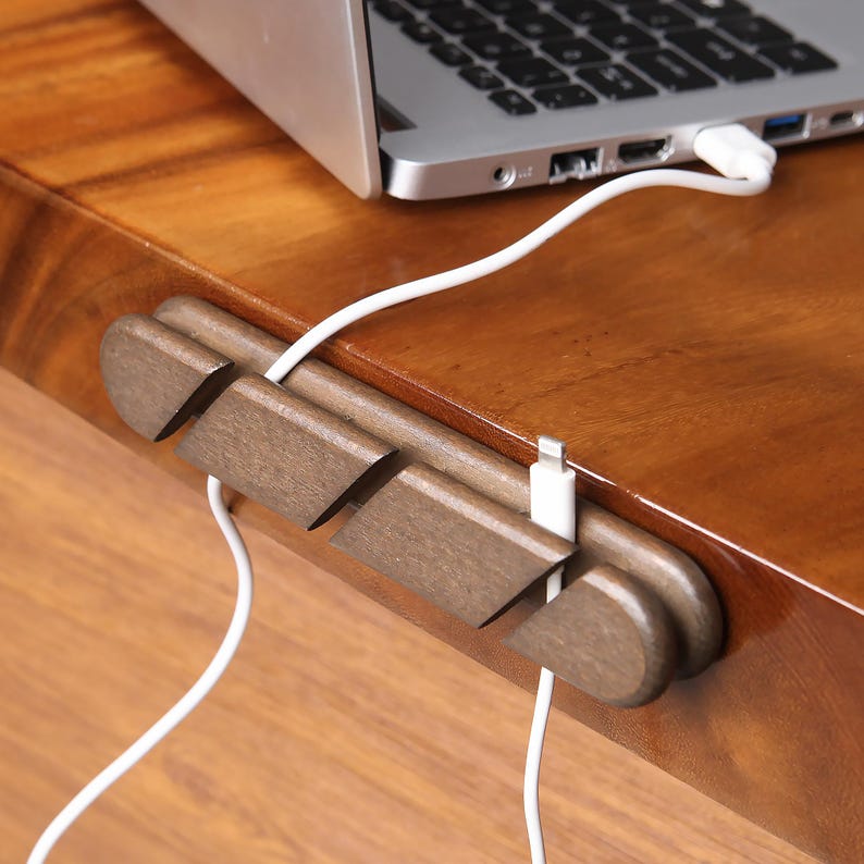 May include: A wooden cable organizer in a warm brown tone, designed to hold and manage charging cables. The organizer is placed on a wooden surface, with a laptop and white charging cables visible.