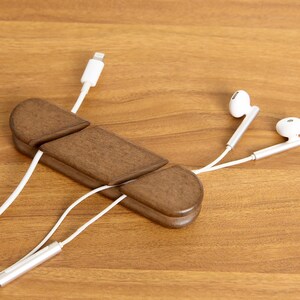 May include: A wooden cable organizer in a warm brown finish, holding white charging cables and earbuds. The organizer has a unique, segmented design. The earbuds and charging cable are white.