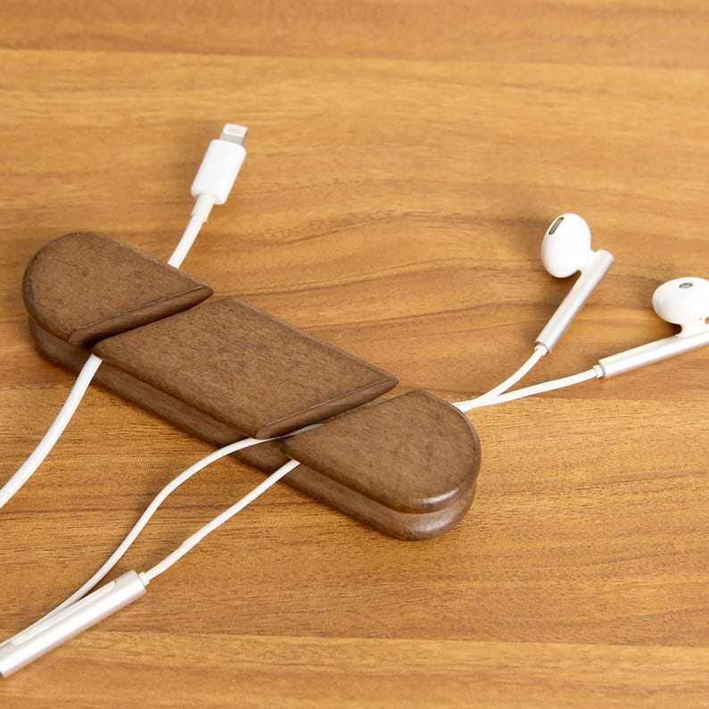 May include: A wooden cable organizer in a warm brown finish, holding white charging cables and earbuds. The organizer has a unique, segmented design. The earbuds and charging cable are white.