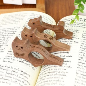 May include: Wooden cat-shaped book holders with engraved details. One features a cat face and open book, another has paw prints, and a third has the name "Mark". The holders are resting on an open book.
