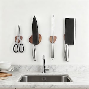 May include: A set of kitchen knives and scissors mounted on a white wall above a stainless steel sink. The knives include a chef's knife, cleaver, and sharpening steel, each held by a wooden magnetic holder. The scissors are also held by a wooden magnetic holder.