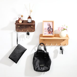 May include: Two wooden wall shelves with hooks, one dark brown and one light brown. The dark brown shelf has the text "Happy Birthday" and holds a small figurine and a bottle. The light brown shelf holds a framed photo and a vase with pink flowers. A black purse and handbag hang below.
