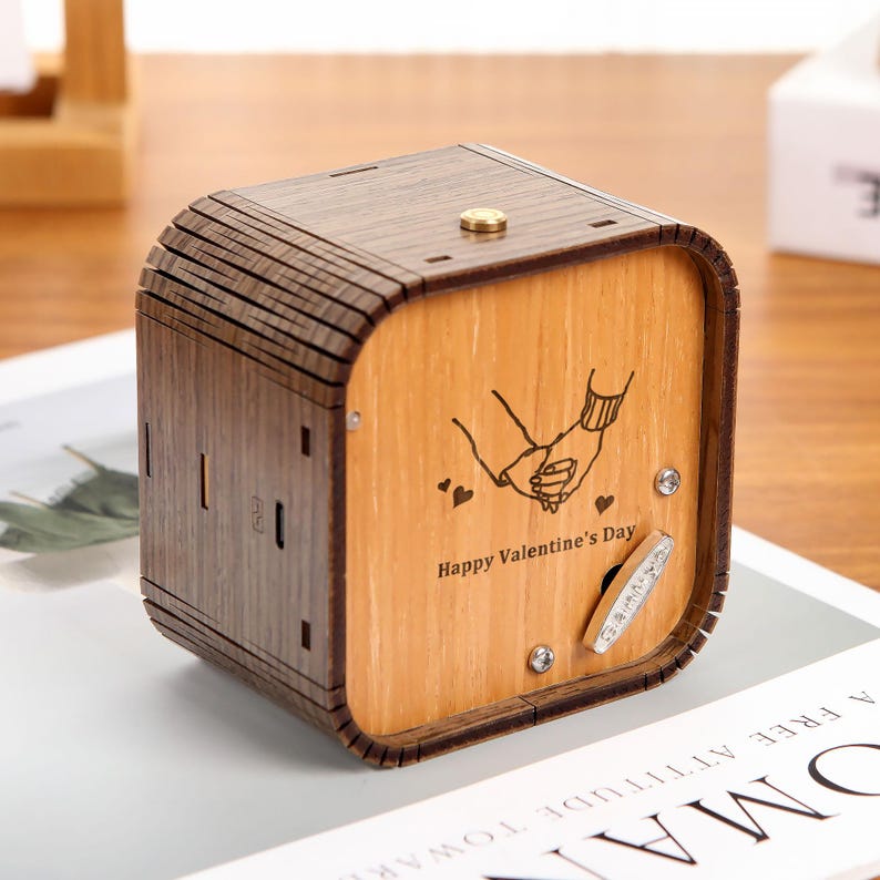 May include: Wooden music box with a light brown finish and a darker brown top. The front features an engraving of two hands holding each other, with the text "Happy Valentine's Day". A silver crank and small silver studs are visible.