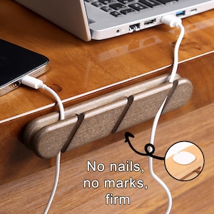 May include: A wooden cable organizer with three slots, holding white charging cables for a laptop and a phone. The organizer is brown and has the text "No nails, no marks, firm".