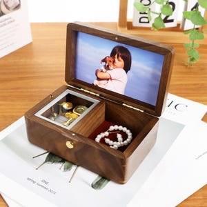 Könnte beinhalten: Eine Holzschmuckschatulle mit Klappdeckel, mit einem Foto einer Person, die einen Hund hält. Die offene Box zeigt Fächer mit Schmuck und einer Perlenkette. Der Innenraum ist mit rotem Stoff ausgekleidet.
