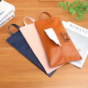 Pode incluir: Três porta-lenços de couro em azul marinho, rosa blush e castanho. O porta-lenços castanho tem uma pega e o texto "Merry Christmas" com um desenho de árvore de Natal. O lenço é visível. Os porta-lenços estão numa superfície de madeira.