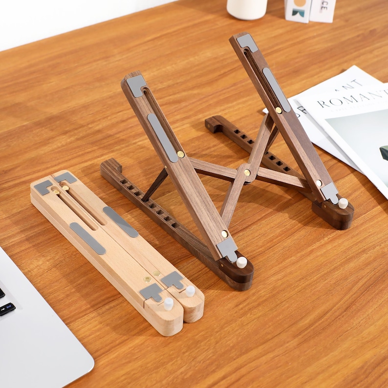 May include: Two wooden laptop stands, one light and one dark, are displayed. They are adjustable and foldable, featuring grey rubber pads. One is assembled, the other folded. The stands are on a wooden surface. The stands are made of wood.
