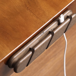 May include: A wooden cable organizer, measuring 1.18 inches long and 0.16 inches wide, is mounted on a wooden surface. A white charging cable is inserted into the organizer. The organizer is a dark brown color.