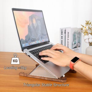 May include: A wooden laptop stand with a triangular stable structure. The stand is holding a silver laptop. The text on the image says "Loading &asymp;5kg" and "Triangular Stable Structure".