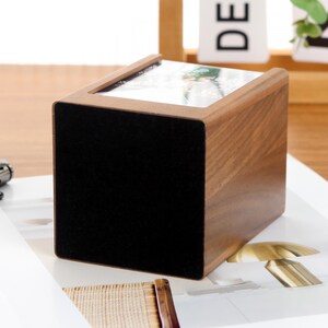 May include: A wooden cube-shaped box with a black front panel and a glass top. The box has a natural wood grain finish and appears to be sitting on a white surface. The top of the box has a picture inside.