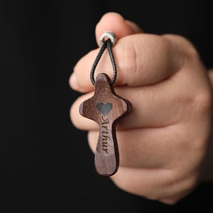 Könnte beinhalten: Dunkelbrauner Holz-Kreuzanhänger mit schwarzem Band. Das Kreuz hat ein herzförmiges Design und den eingravierten Namen "Arthur". Der Anhänger wird vor einem dunklen Hintergrund gehalten.