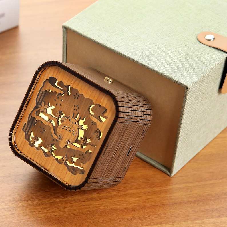 May include: A wooden music box with a laser-cut design featuring a cartoon character and natural elements. The box is square-shaped with a hinged lid and sits next to a green gift box. The music box is made of light and dark brown wood.