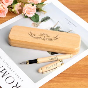 May include: A light-colored wooden pen case and two pens. The case is rectangular with the word "wedding" and "Lorem Ipsum" engraved on the lid. One pen is labeled "Amily" and the other "25.12.25".