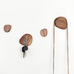 May include: Four pebble-shaped wooden wall hooks. The hooks are made of warm-toned wood and are mounted on a white wall. One hook holds keys, and another holds a gold chain. Ideal for a hallway or entryway.