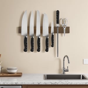 Puede incluir: Un conjunto de cuchillos de cocina con mangos negros y con vetas de madera, expuestos en una tira magnética de madera. El juego incluye cuchillos de varios tamaños, una chaira y unas tijeras de cocina. Los cuchillos están montados en una pared beige.