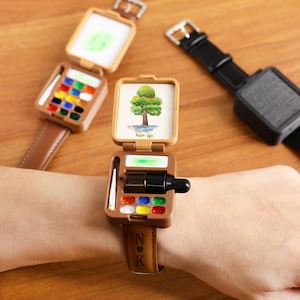 May include: Brown leather watch with a distinctive design. The watch face opens to show a watercolour palette and a miniature painting of a tree with the words "hope life". The watch strap is engraved with the initials "N K".
