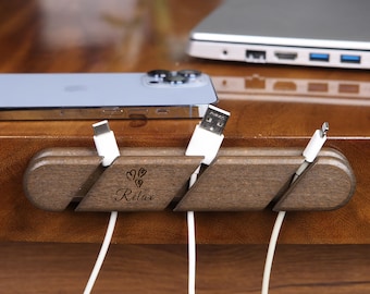 May include: A wooden cable organizer with the word "Relax" and heart-shaped designs. The organizer holds several white charging cables, with a phone and laptop visible in the background. The organizer is made of dark brown wood.