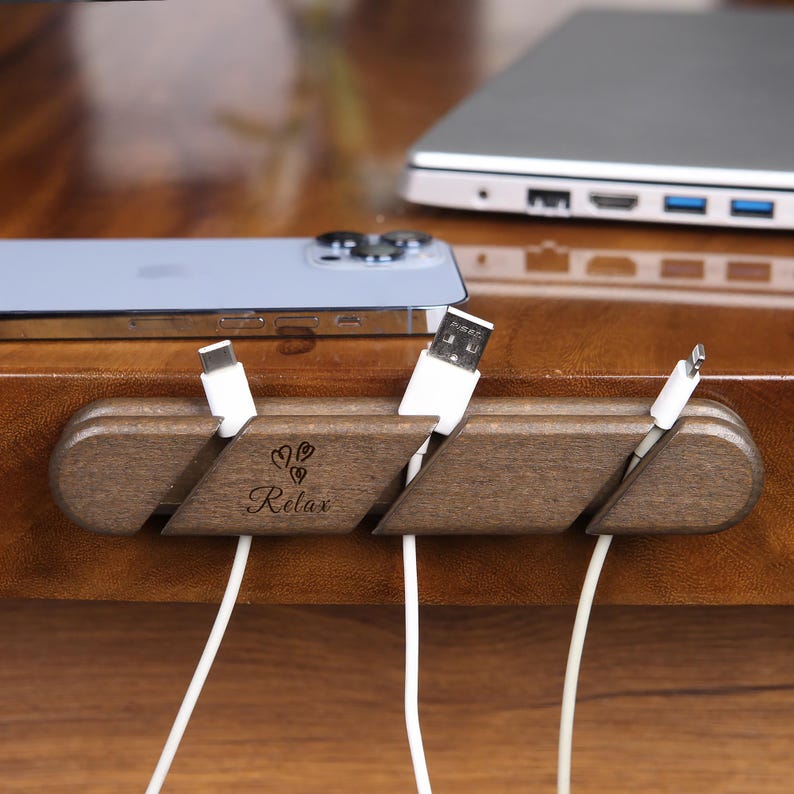 May include: A wooden cable organizer with the word "Relax" and heart-shaped designs. The organizer holds several white charging cables, with a phone and laptop visible in the background. The organizer is made of dark brown wood.