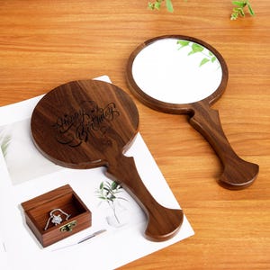 Może przedstawiać: Dwa drewniane lusterka do rąk z ciemnobrązowym wykończeniem. Jedno lusterko ma okrągłą powierzchnię odbijającą światło, a na drugim wygrawerowano napis "Happy Birthday". Widoczne jest również małe drewniane pudełko na biżuterię.