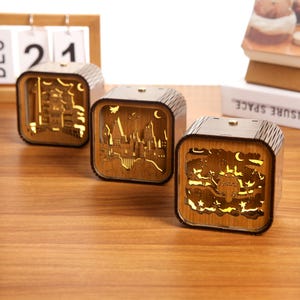 May include: Three wooden, cube-shaped night lights with intricate cut-out designs. Each light features a different scene, including a castle, a cityscape, and a whimsical creature. The lights are illuminated from within, creating a warm, inviting glow.