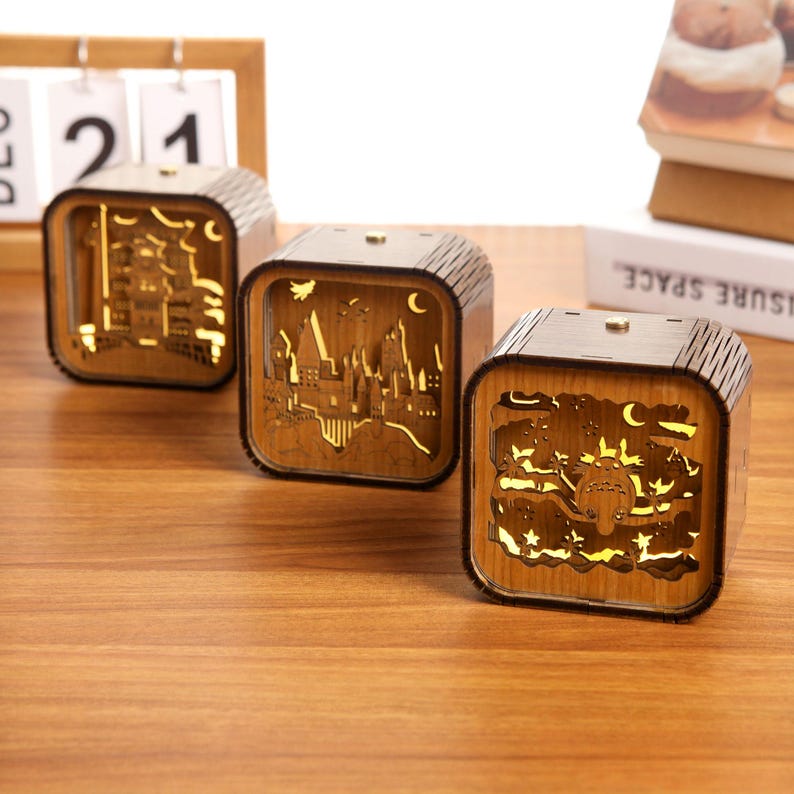 May include: Three wooden, cube-shaped night lights with intricate cut-out designs. Each light features a different scene, including a castle, a cityscape, and a whimsical creature. The lights are illuminated from within, creating a warm, inviting glow.