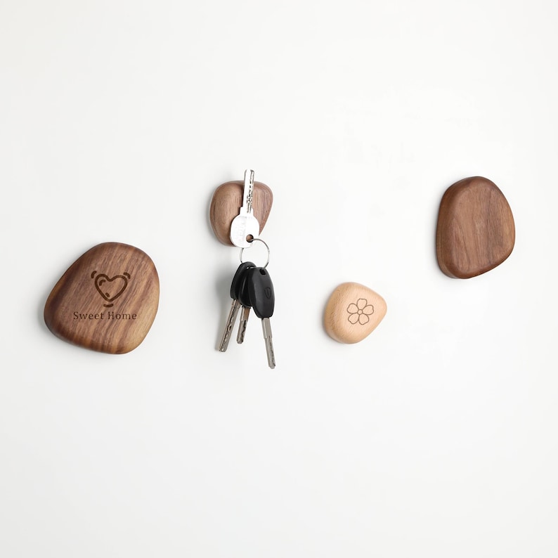 May include: Wooden key holders in the shape of smooth stones. One holder has a heart and the words "Sweet Home" engraved. Another holds keys. A third has a flower design. The key holders are various shades of brown.