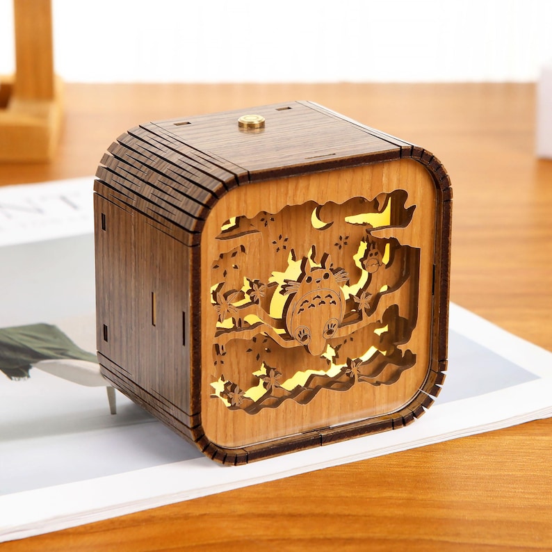 May include: A wooden cube-shaped night light with a detailed cutout design. The design features a cartoon character, trees, and stars. The light glows from within, creating a warm ambiance. The night light is made of light brown wood.