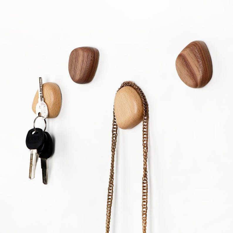May include: Wooden wall hooks in various shades of brown. One hook holds keys, another a gold chain. The hooks have a smooth, organic shape, and are mounted on a white wall. The hooks are made of wood.