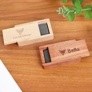 Puede incluir: Dos cajas de madera con tapas correderas, una de color claro y otra de color marrón rojizo. Cada caja contiene una piedra porosa de color gris oscuro. La caja clara está grabada con un diseño de corazón y las palabras "Love & Always". La otra caja está grabada con un corazón y el nombre "Bella".