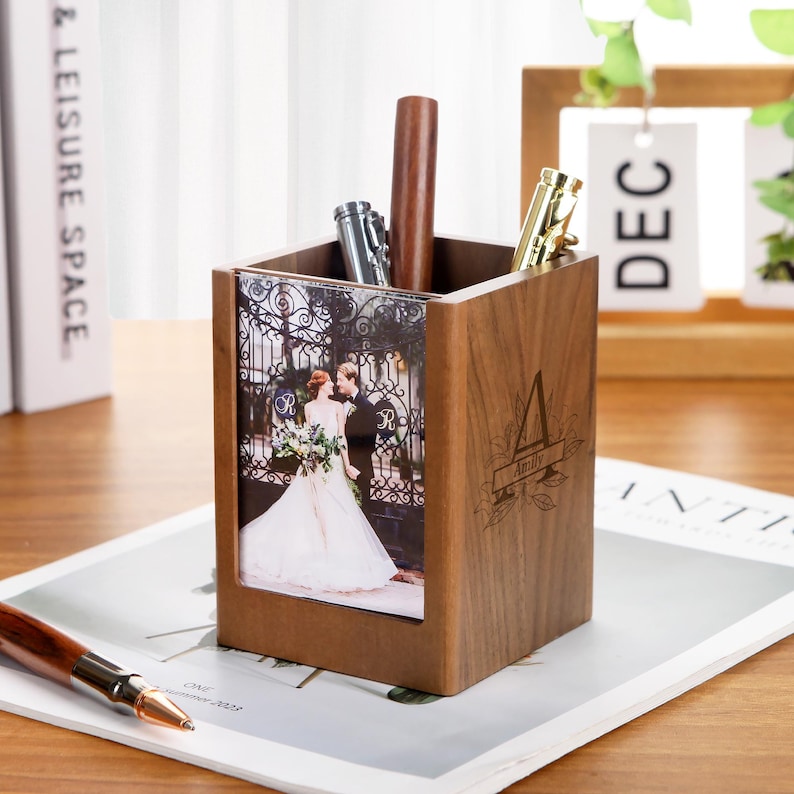 May include: A wooden pen holder with a photo of a couple in front of a wrought iron gate. The pen holder has a personalized initial and name. Several pens are inside the holder, including a silver pen and a gold pen.