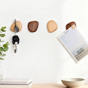 May include: Wooden pebble-shaped wall hooks in varying shades of brown and tan. One hook holds keys, another is empty, and a third holds a paper with a picture of a van. The hooks are mounted on a white wall.