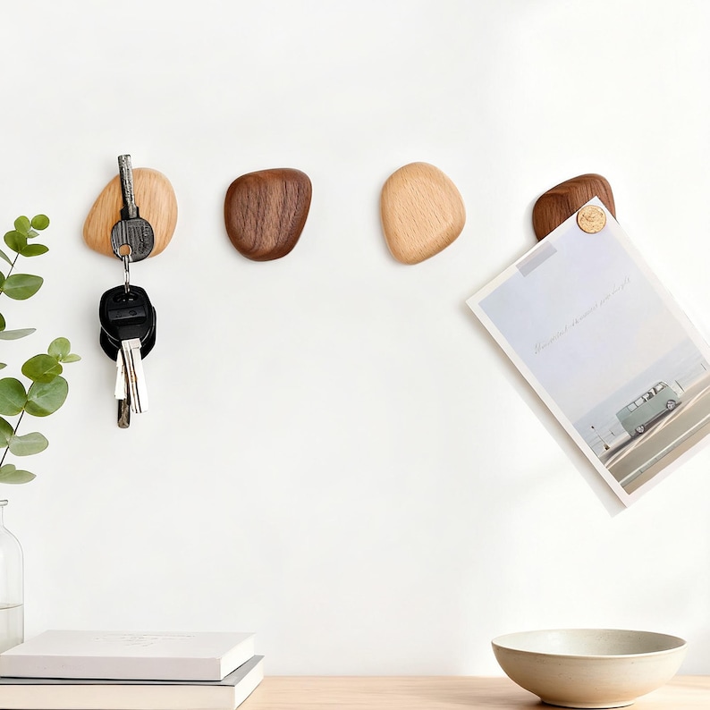 May include: Wooden pebble-shaped wall hooks in varying shades of brown and tan. One hook holds keys, another is empty, and a third holds a paper with a picture of a van. The hooks are mounted on a white wall.