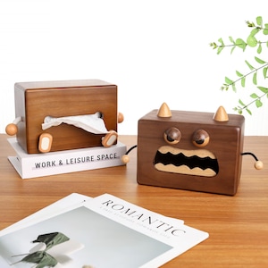 May include: Two wooden tissue box holders with cartoon monster designs. One is light brown with a rectangular shape, and the other is dark brown with a square shape. The light brown box has the text "WORK & LEISURE SPACE" on the book it is sitting on.