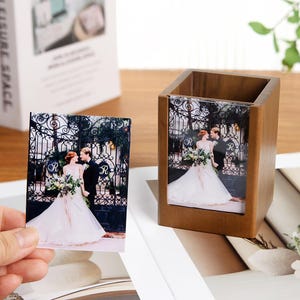 May include: A wooden pen holder with a photo of a couple in wedding attire. A separate photo of the same image is held in a hand. The photos feature a bride and groom in front of a wrought iron gate.