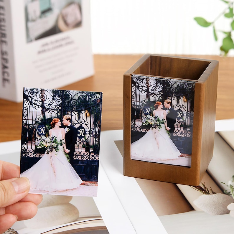 May include: A wooden pen holder with a photo of a couple in wedding attire. A separate photo of the same image is held in a hand. The photos feature a bride and groom in front of a wrought iron gate.