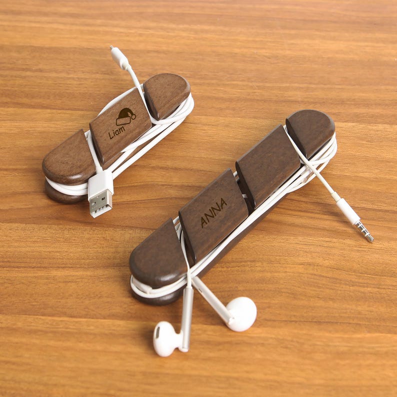 May include: Two wooden cable organizers with white cords. One has "Liam" engraved with a Santa hat, the other says "ANNA". The organizers are brown and have a slot to hold the cable in place. The background is a wooden surface.