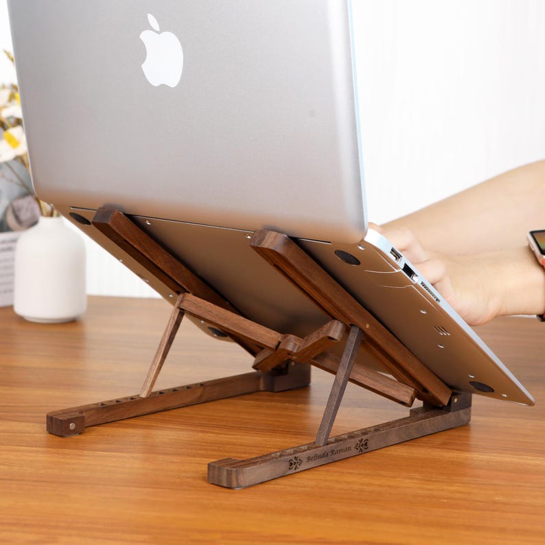 May include: A dark brown wooden laptop stand supporting a silver laptop. The stand is adjustable and made of wood. The laptop has the Apple logo. The stand is on a wooden surface.