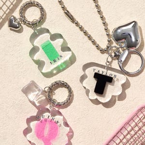May include: Three personalized keychains with silver-tone hardware. One keychain has a heart charm and a green design with the name "IRIS". Another has a heart charm and the name "TAYLOR". The third has a clear charm with the name "QUINN".