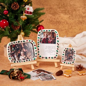 May include: Three white ceramic picture frames with colorful accents, each holding a photograph, displayed on small wooden easels. A Christmas tree with ornaments is in the background, along with additional holiday decorations.