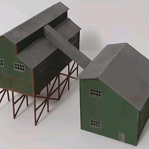 May include: A model train building kit with two structures, one with a raised platform and a slanted roof, and the other with a flat roof. Both buildings are green with gray roofs and windows.