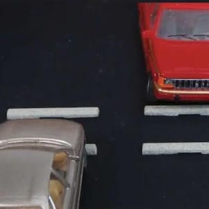 May include: A silver car and a red pickup truck are parked, with gray concrete parking stops in front of them. The vehicles are on a black surface. The red truck has a chrome grill.