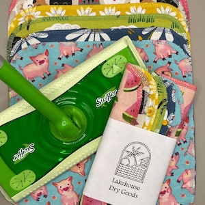 May include: A green Swiffer mop and a stack of colorful, patterned cleaning cloths. The cloths feature designs of pigs, cats, and watermelons. A label reads "Lakehouse Dry Goods".