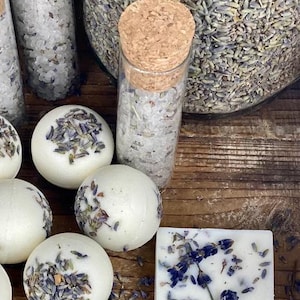 Könnte beinhalten: Eine Sammlung von Lavendel-Badeprodukten, darunter Badebomben, Seifenstücke und Badesalze. Die Badebomben und Seifenstücke sind weiß mit eingebetteten getrockneten Lavendelblüten. Die Badesalze befinden sich in Glasbehältern mit Korkverschlüssen.